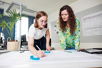 Larina absolviert ihre Ausbildung bei der Zentralen Technik und zeichnet unter Aufsicht einer ehemaligen dualen Studentin etwas auf einem Bauplan ein.
