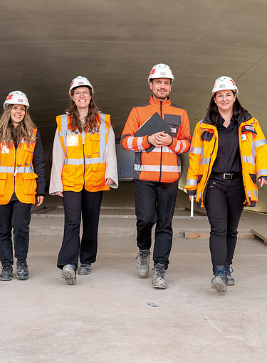 Das Foto zeigt vier Mitarbeiter:innen der Zentralen Technik, die auf der Baustelle mit Sicherheitskleidung durch einen Tunnel laufen.