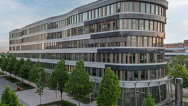 Das Bild zeigt ein Gebäude mit einer runden Fassade, vielen Fenstern und Bäumen davor.