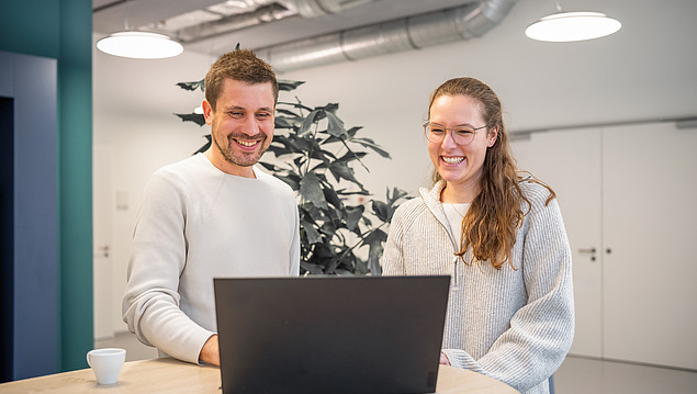 The picture shows two employees of the Zentrale Technik smiling as they look at a laptop and talk about the HOAI service phases.