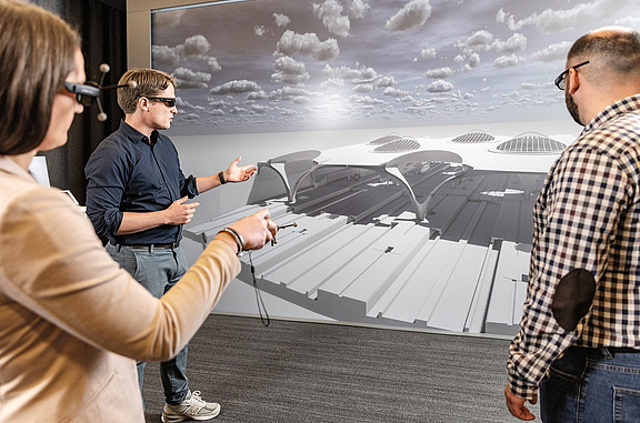 Three people view a 3D model on a large screen using BIM technology and VR glasses.
