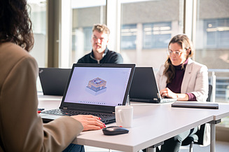 Personen arbeiten gemeinsam an Laptops in einem Besprechungsraum, während auf einem Bildschirm ein Gebäudemodell zu sehen ist - eine typische Arbeitsumgebung für ganzheitliche Planung im Generalplaner Team.
