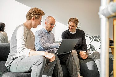 Nachhaltigkeit in der Zentralen Technik. Drei Mitarbeitende sitzen auf einem Sofa und schauen gemeinsam auf einen Laptop, während sie sich über ein Projekt austauschen.