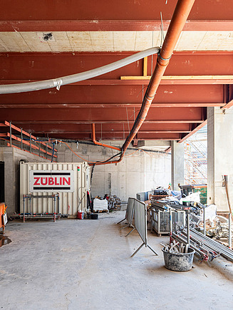 A Züblin site container and building materials are standing in the shadow of a structural slab.