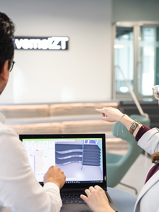 Das Bild zeigt zwei internationale Mitarbeitende, die ein Planungsmodell mit dem fertigen Bauwerk innerhalb eines Gebäudes der Zentralen Technik vergleichen. Im Hintergrund leuchtet das Schild mit der Aufschrift „vernetzt“.