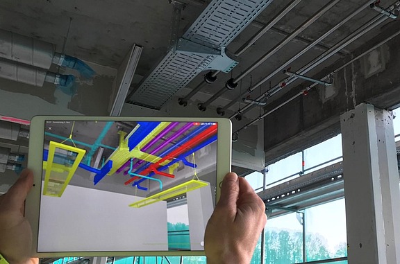 Tablet shows a color 3D model of technical systems in a building, while the real installation of pipes and ducts on the ceiling is visible in the background.