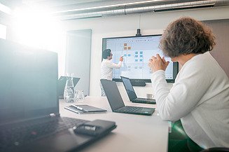Das Bild zeigt zwei Personen in einem modernen Besprechungsraum. Eine Person steht vorne am Bildschirm und präsentiert etwas – Zentrale Technik, Generalplanung
