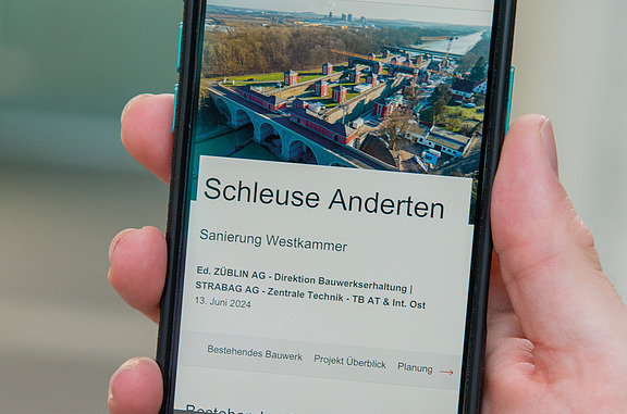 Auf dem Bild ist eine Hand zu sehen, die ein Handy hält. Auf dem Handy ist die Schleuse Anderten zu sehen.