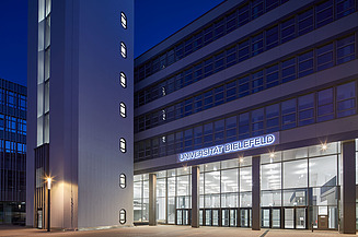 Eingangsbereich des UHG Bielefeld bei Dämmerung mit beleuchteter Glasfassade.