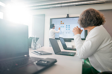 The picture shows two people in a meeting room. One person is standing at the front of the screen and showing something