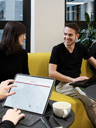 The picture shows four people on a sofa talking. In the foreground you can see a tablet on which a BIM model can be seen