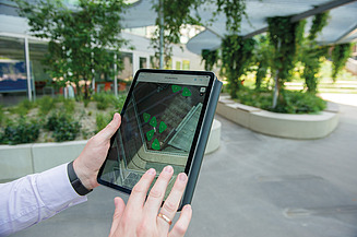 Das Bild zeigt ein Tablet mit einem Kartenausschnitt darauf, der auch im Hintergrund in echt zu sehen ist und symbolisiert damit die Digitalisierung von Standortkarten - GIS in der Zentralen Technik.