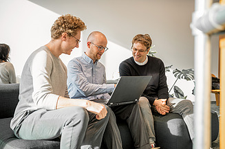 Green Services in der Zentralen Technik. Drei Mitarbeitende sitzen auf einem Sofa und schauen gemeinsam auf einen Laptop, während sie sich über ein Projekt austauschen.