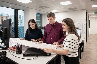 Eine Auszubildende bespricht Konstrukteurspläne mit ihren Arbeitskolleg:innen.