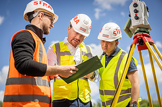 Three STRABAG construction engineers from the Zentrale Technik department stand at a tachymeter.