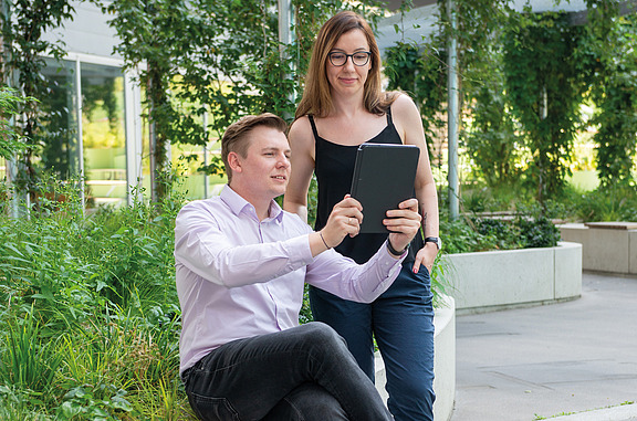 Zwei Personen nutzen im Außenbereich ein Tablet zur gemeinsamen Ansicht von Geodaten, umgeben von moderner Architektur und viel Grün.
