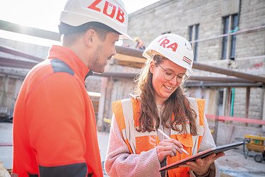 Two employees talking on a construction site; an employee with a tablet shows information, both are wearing hard hats and protective clothing.