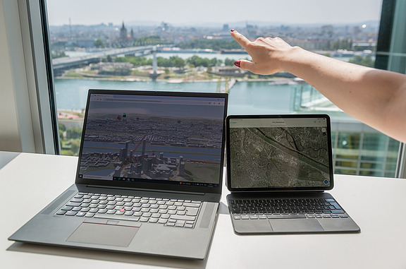 Laptop und Tablet mit geöffneten Geoinformationssystemen vor einem Fenster mit Blick auf eine Stadtlandschaft, während eine Person auf die Umgebung zeigt.