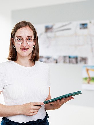Larina, a draughtswoman in training at Zentrale Technik, stands in her office with a tablet in her hand.