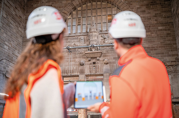 Auf dem Bild sieht man zwei Personen in Baustellenkleidung auf der Baustelle, die ein Tablet halten.