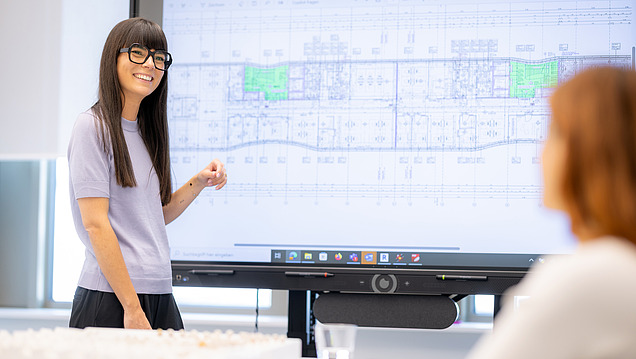 An employee from Zentrale Technik department stands in front of a large screen with a plan of the service phases.