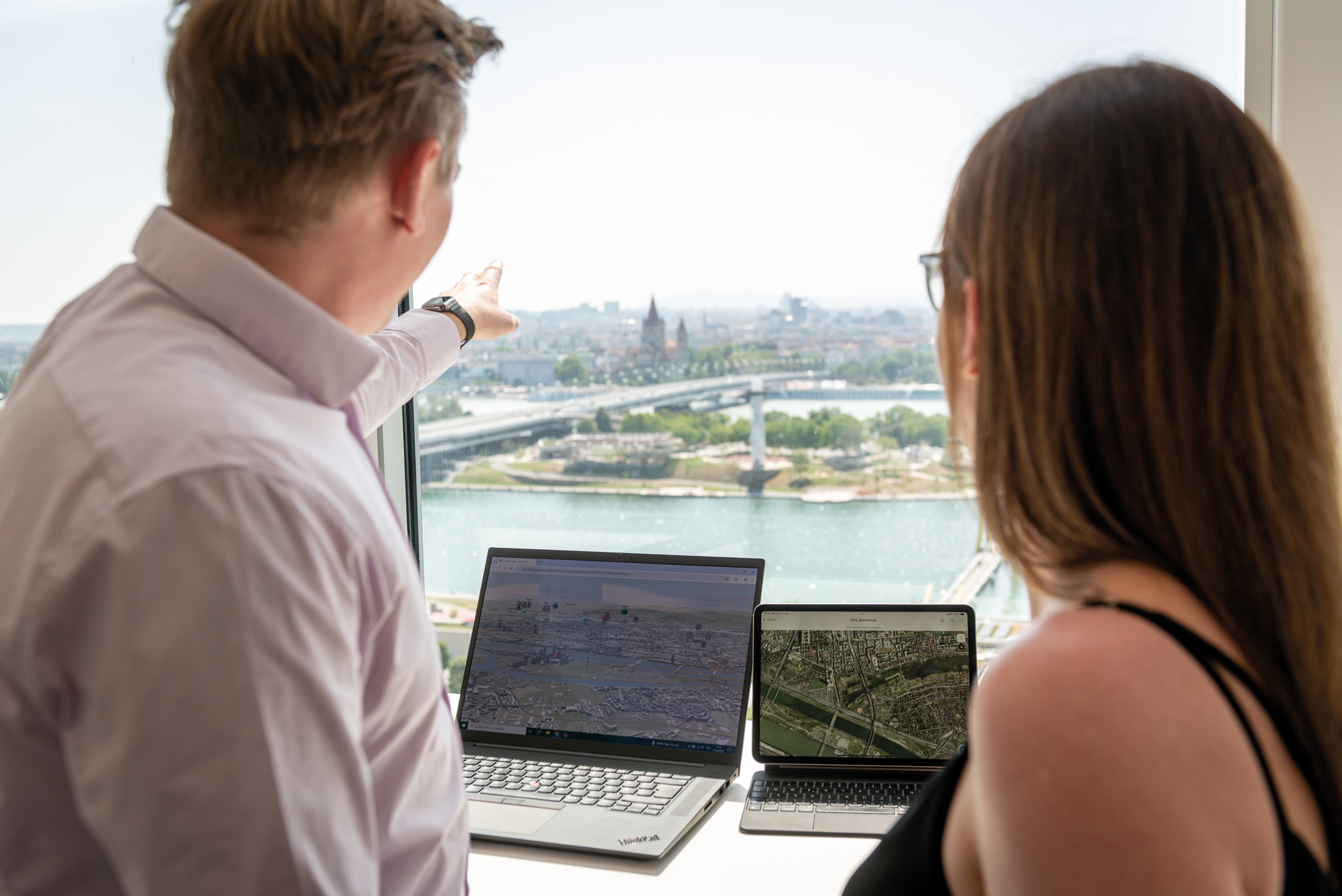 Zwei Personen betrachten auf ihren Laptops Geodaten vor einem großen Fenster mit Blick auf eine Stadtlandschaft.