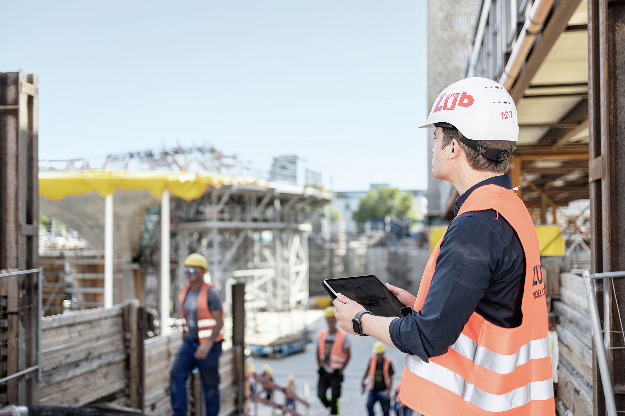 A person stands on a construction site holding a tablet with a BIM model on it
