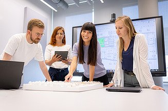 Ein Planungsteam blickt mit einem Trainee auf ein Architekturmodell, das in einem Besprechungsraum auf einem Tisch steht.
