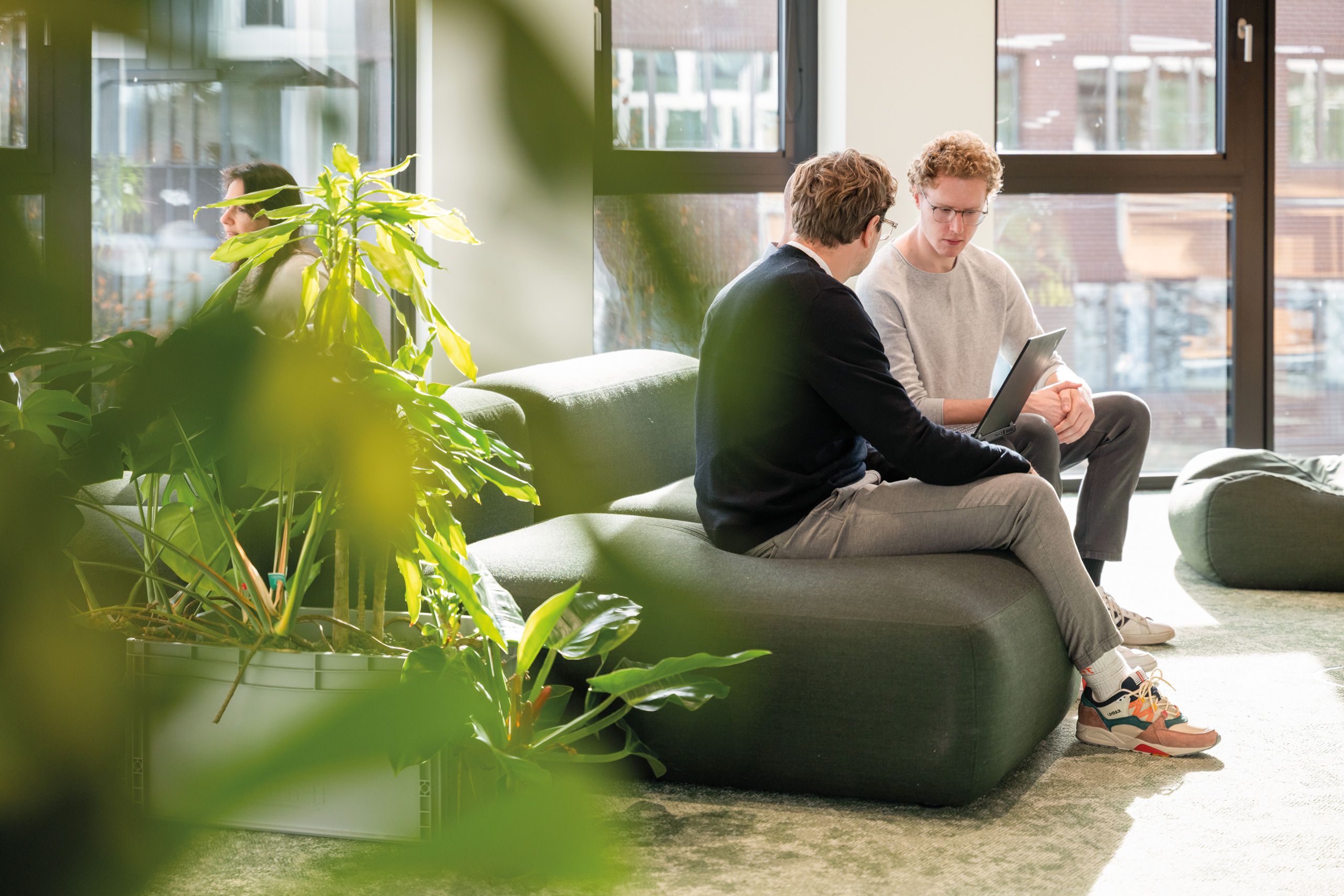 Two people are sitting on green seating furniture in a modern lounge having a conversation. Next to them are large green plants, and daylight floods into the room through the large windows