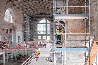 Ein Sinnbild für Bauen im Bestand: Im Vordergrund bearbeitet ein Bauarbeiter auf einem Baugerüst die denkmalgeschützte Fassade, im Hintergrund ist der Rohbau der neuen Zwischenebene zu erkennen.               