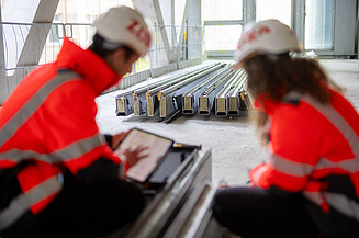 In the foreground, two civil engineers are looking at a tablet; prefabricated components are lying in the background.