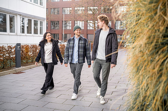 Three people walk together on a paved path in front of office buildings, surrounded by hedges and plants.