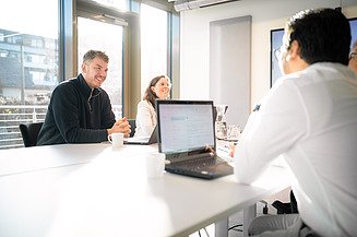 Cheerful employees from Zentrale Technik sit in a meeting room lit by the morning sun.