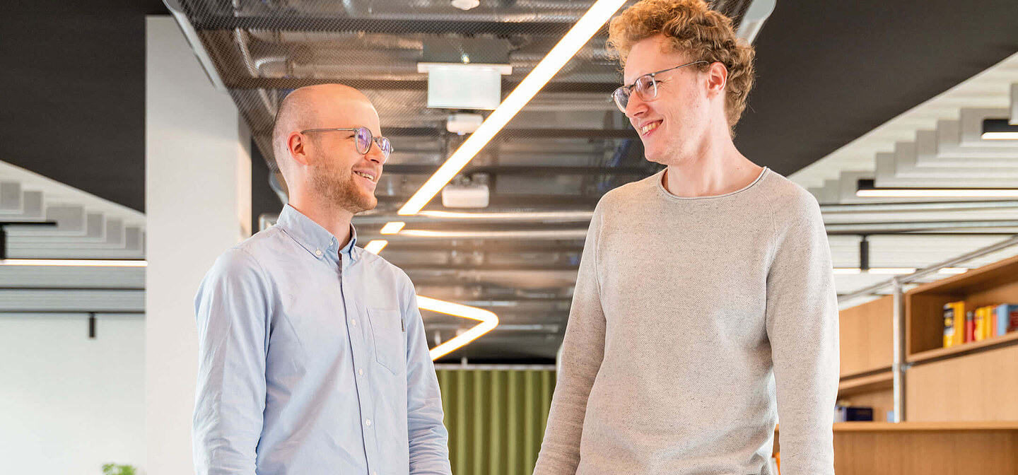 Zwei Mitarbeiter der Zentralen Technik stehen im Büro, reden über ihre Karriere und halten ihre Sicherheitsweste und ihren Sicherheitshelm in der Hand.