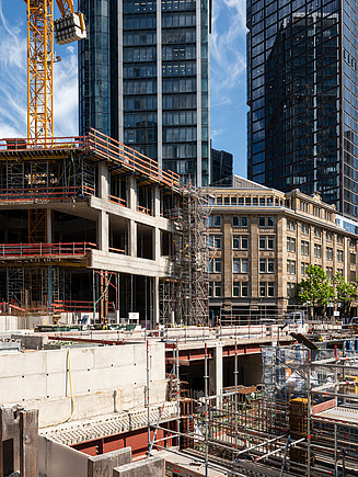 In the foreground, a concrete mixer is being lifted into the air by a crane; in the background, construction workers are at work on the CBT Frankfurt building site.